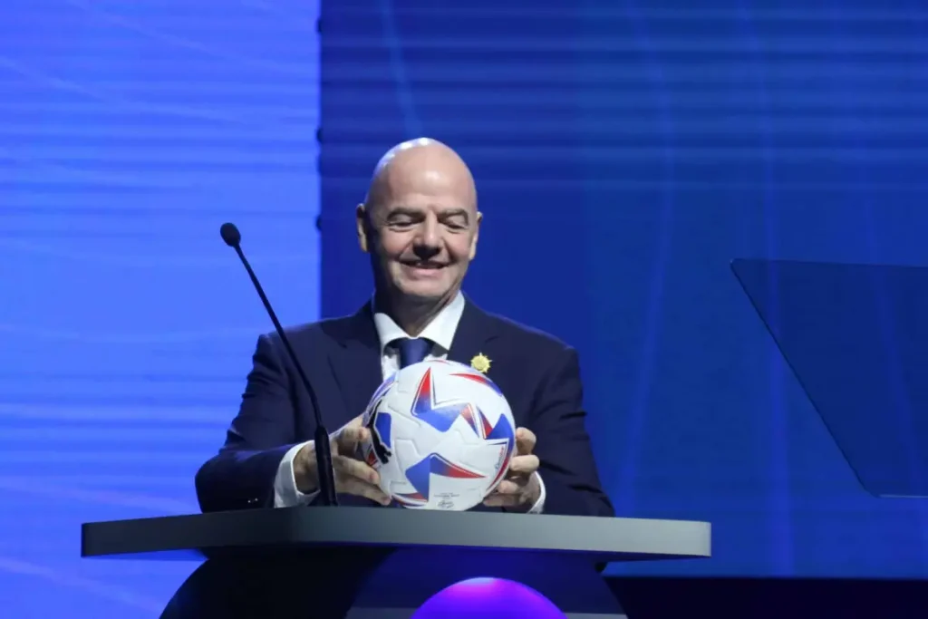 Infantino habría negociado la final del Mundial 2030 para asegurar su continuidad en la FIFA 1 Gianni infantino sosteniendo un balon en un podio y vistiendo un traje negro con camisa blanca.